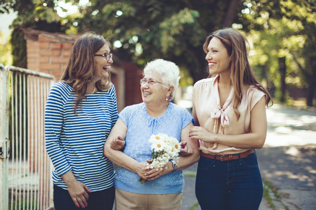 Three women, one elderly and two younger, smiling and walking outside, the elderly woman holds flowers.