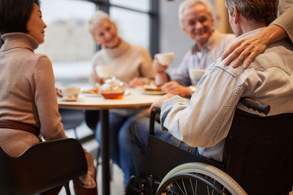 Four people enjoying tea at a table, with one person in a wheelchair and another with a hand on his shoulder.