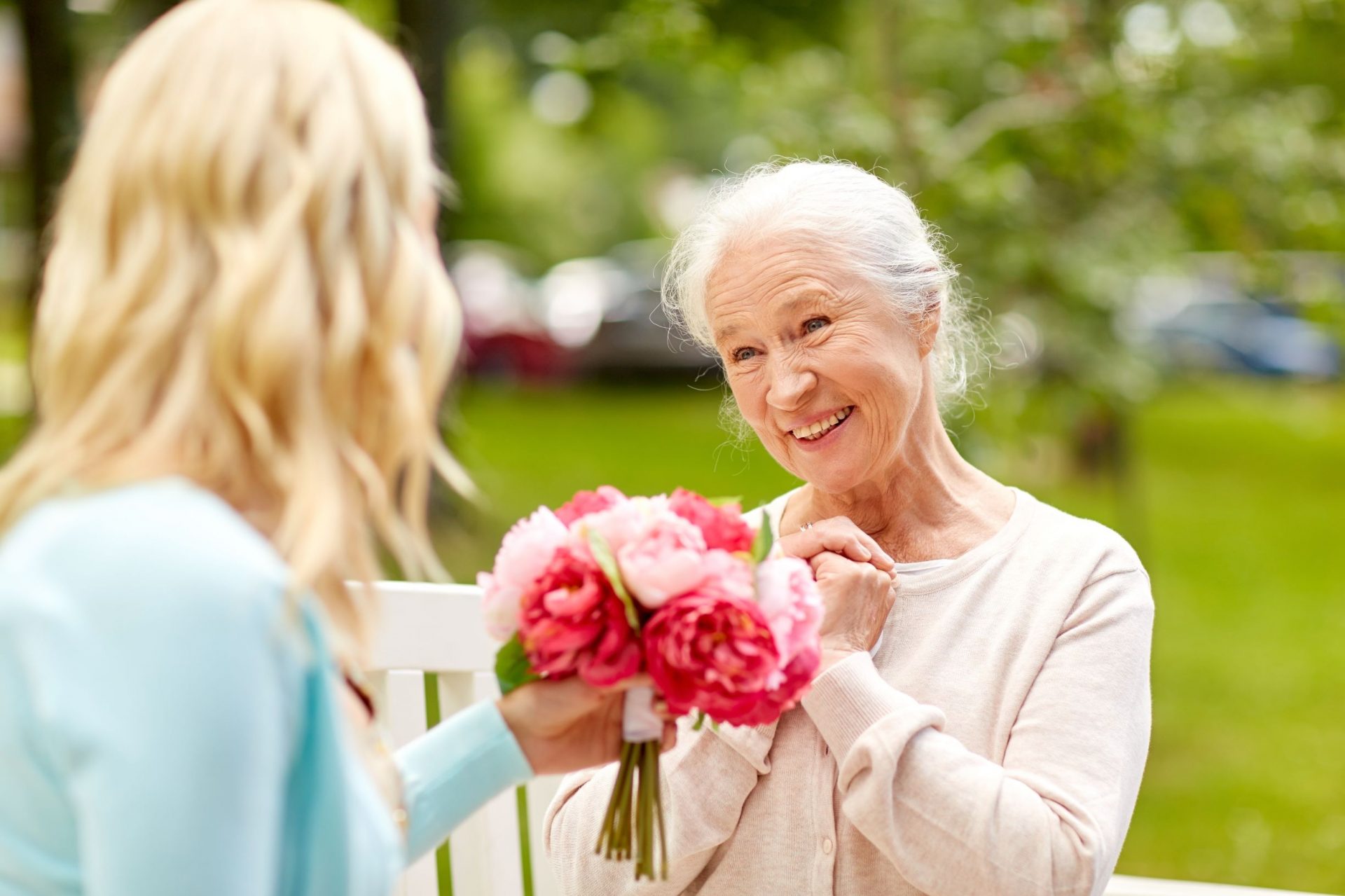 Elderly woman on a bench, smiling as younger woman hands her a bouquet of flowers outdoors.