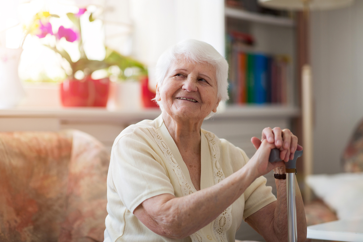 Elderly woman with white hair sitting in a cozy room, holding a cane, and smiling warmly.