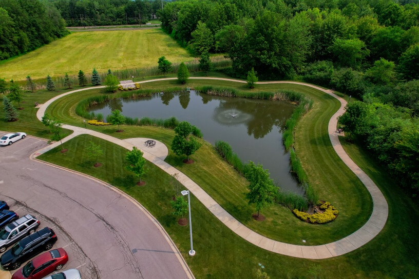 Aerial view of a small pond surrounded by a curved walkway, trees, and parked cars in a green area.