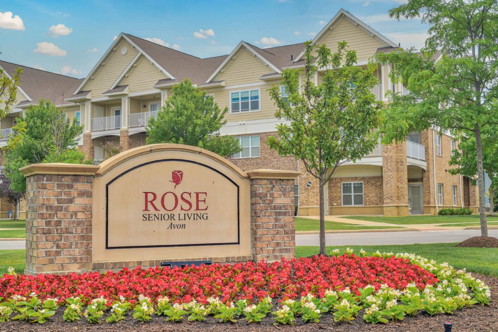 Entrance sign reading "Rose Senior Living Avon" with landscaped flowers and buildings in the background.