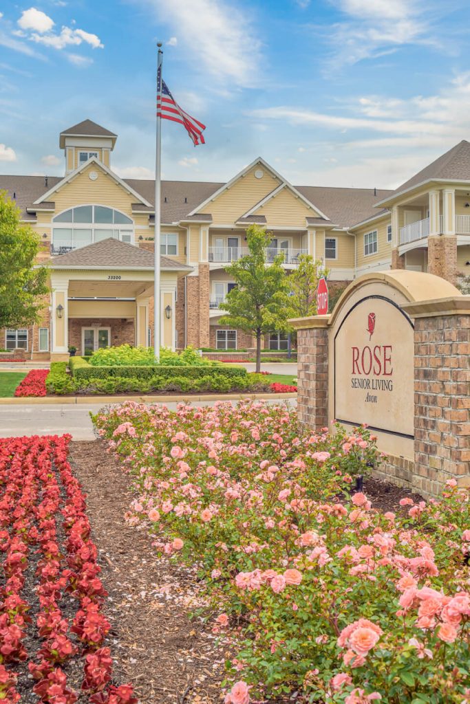 Rose Senior Living building with manicured gardens, American flag, blue sky, and blooming flowers.