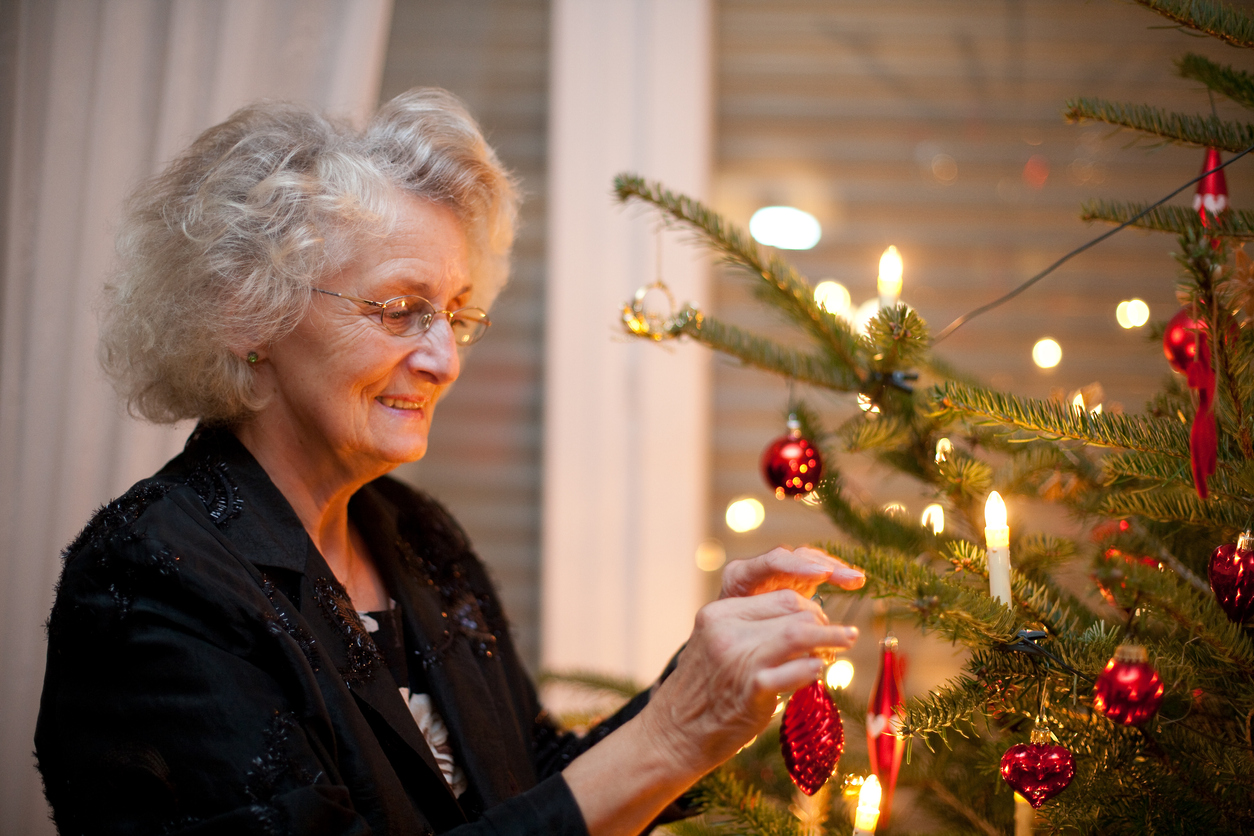 Elderly woman in glasses decorating a Christmas tree with red ornaments and candles.