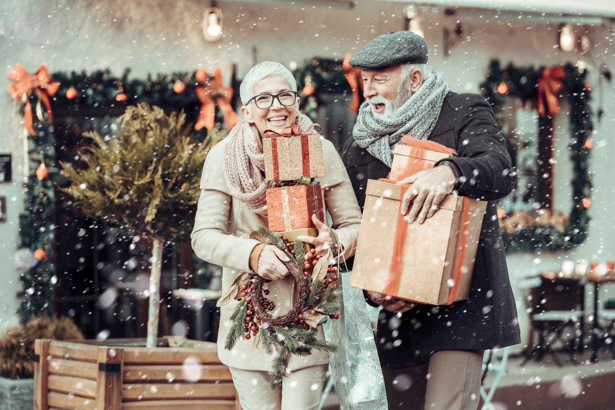 Elderly couple smiling while carrying Christmas gifts and a wreath in a snowy street