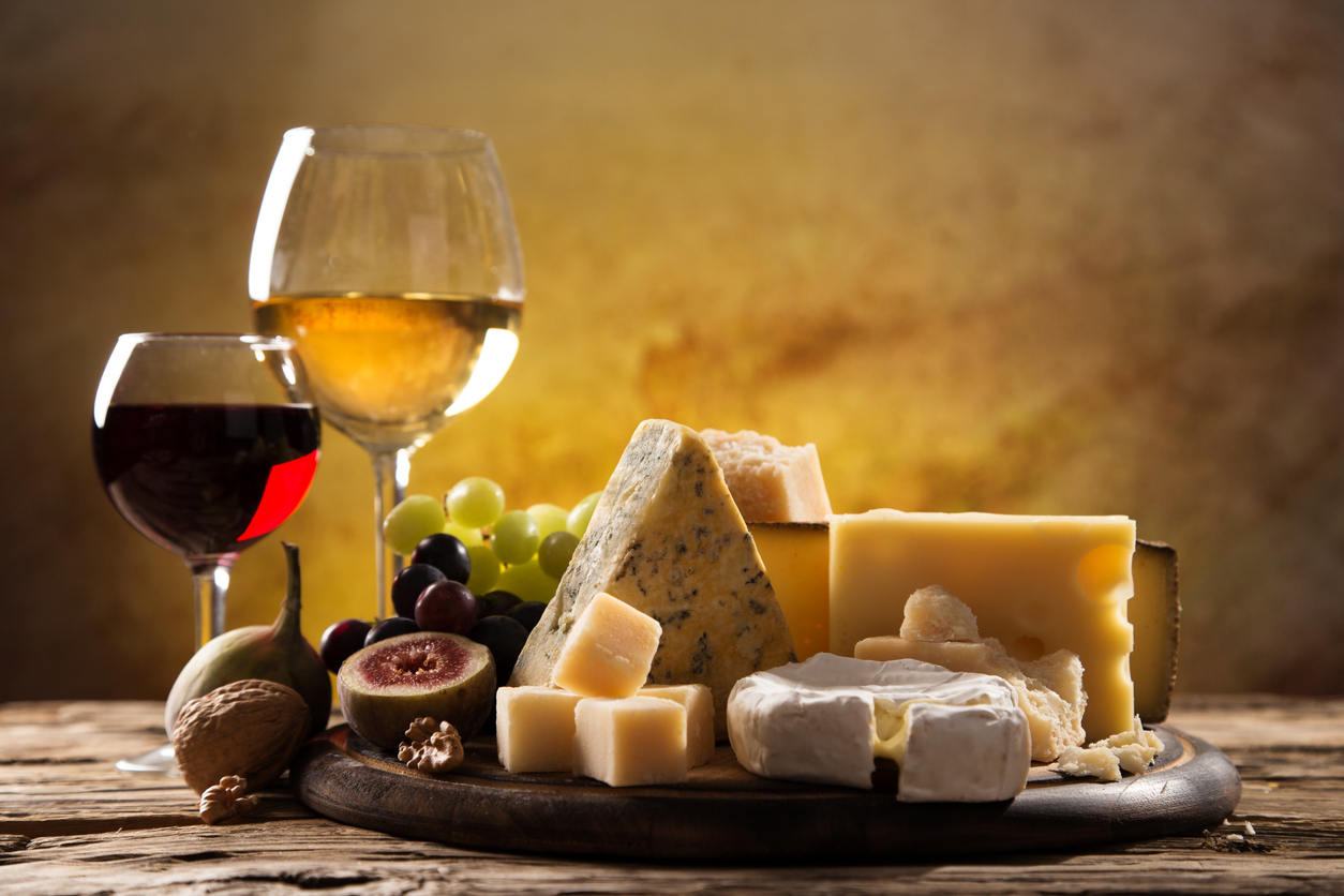 Various cheeses and fruits on a wooden board with glasses of red and white wine in the background.